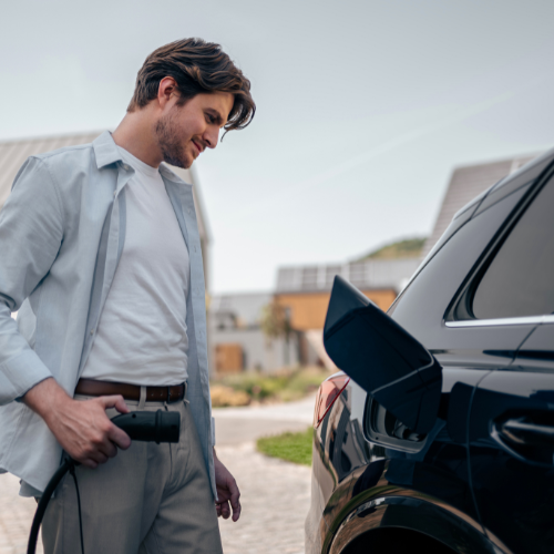 A casually dressed man on a sunny day, holding an EV charging cable and standing next to a black electric car, indicating that he is about to charge his car.