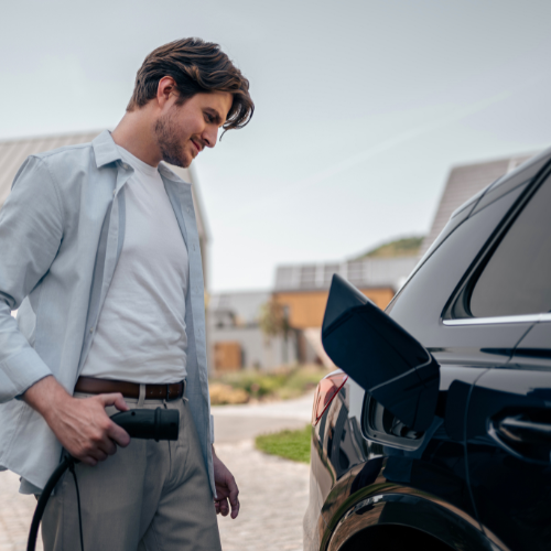 A man wearing a casual outfit standing next to his car on his driveway with a charging cable in hand.