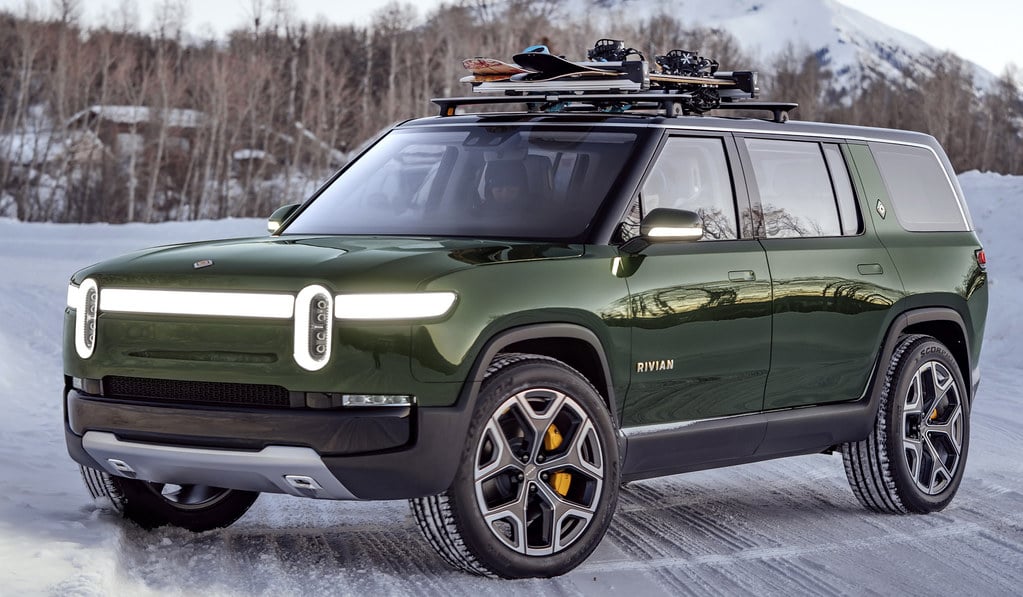 A picture of an army-green Rivian RS1 EV SUV parked dynamically in a snowy landscape with its lights still on.