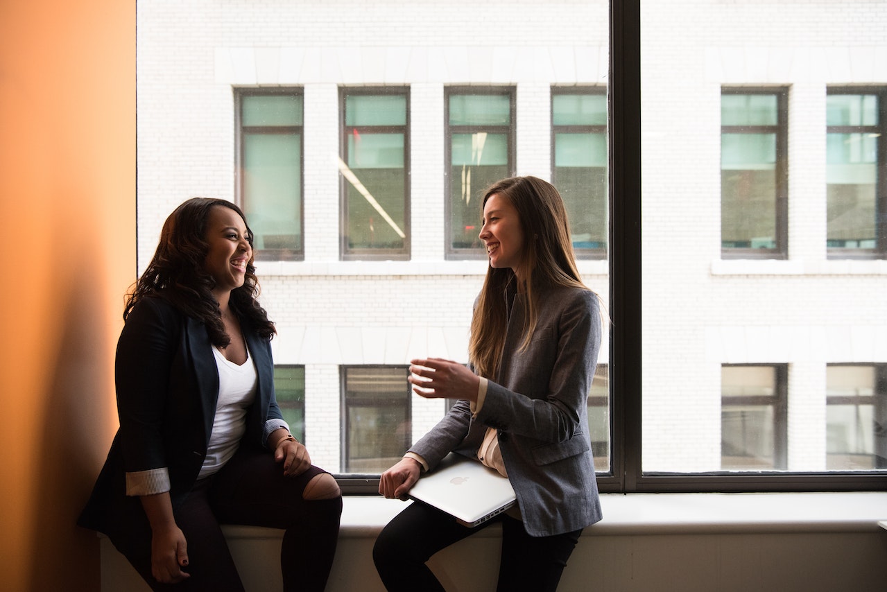 Two women in smart casual attire sitting inside on a window sill chatting to each other