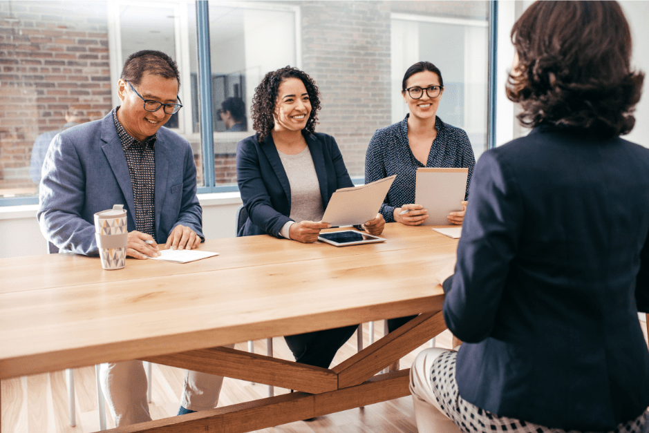 A woman facing three interviewers, the interviewer in the middle is smiling at her whilst the other also smile and look at her resumé