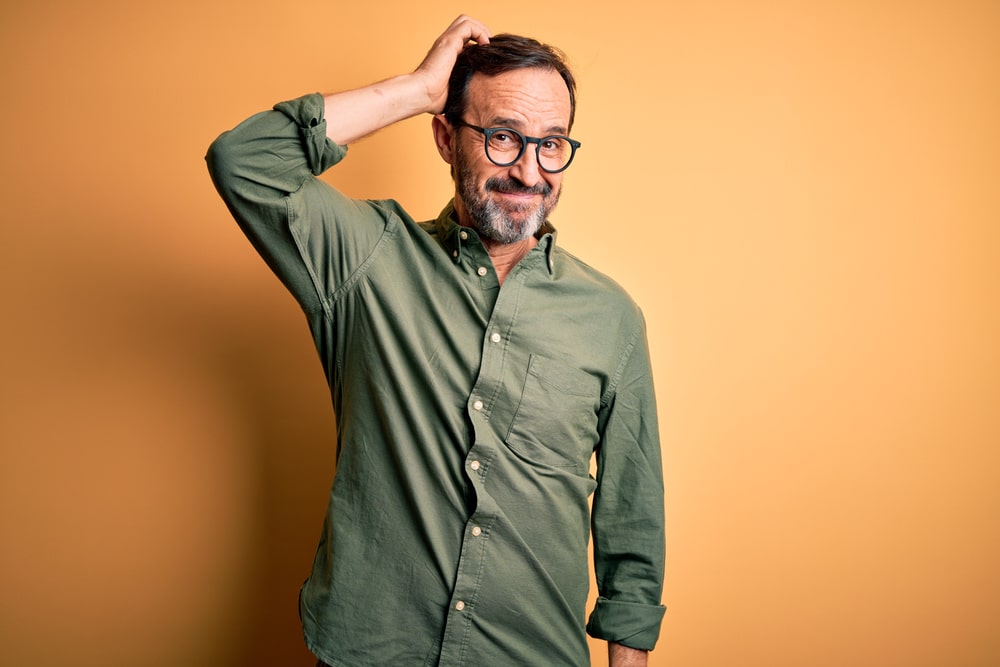 A middle-aged man wearing a casual green shirt and modern glasses looking doubtful in front of an orange background.