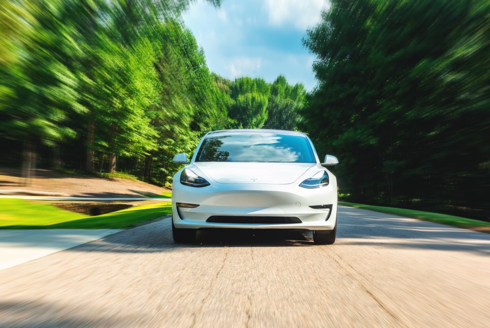 White Tesla model 3 driving down road surrounded by trees.