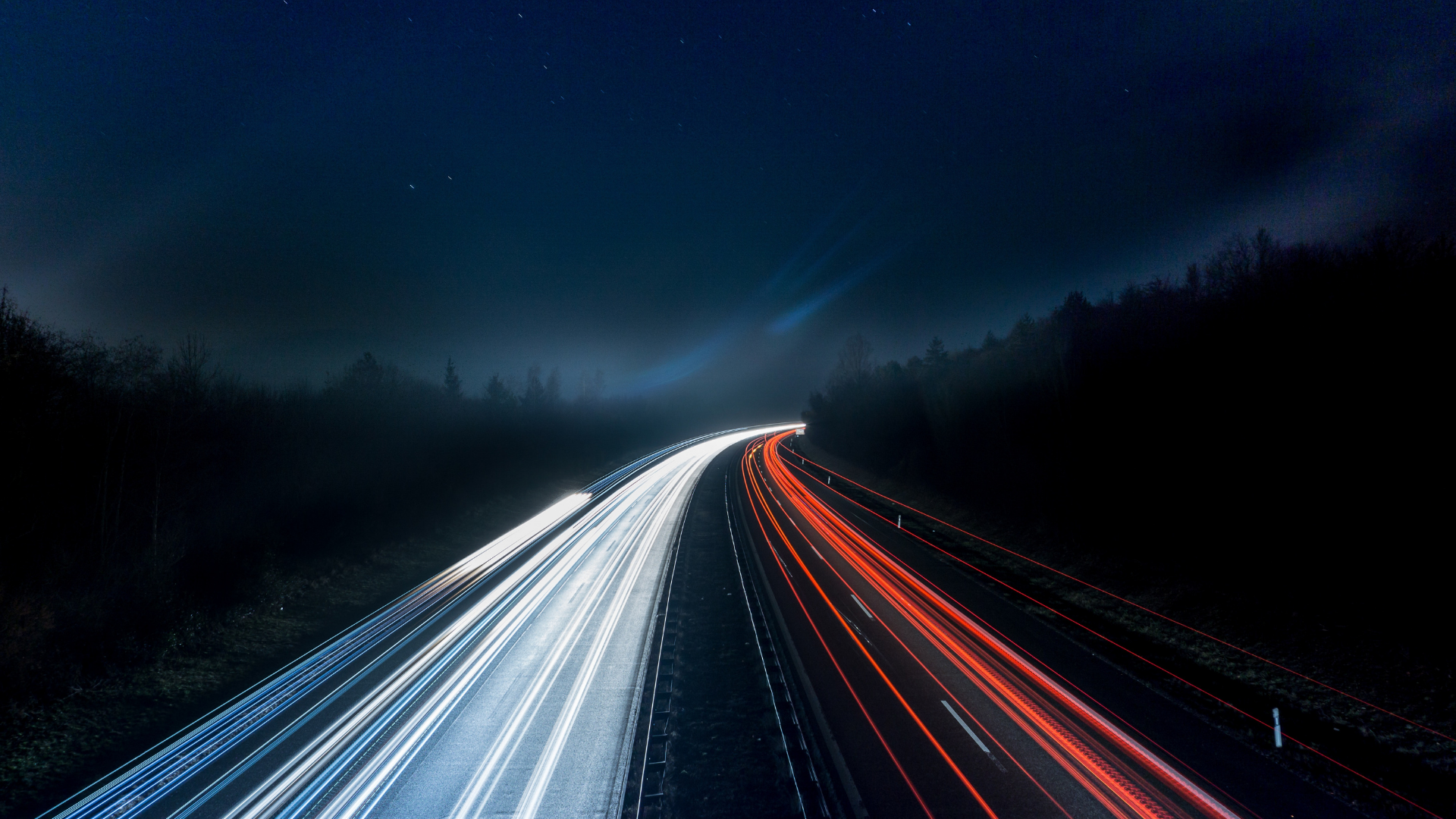 Ein Foto einer belebten Autobahn bei Nacht, aufgenommen mit einer langen Verschlusszeit, um die weißen und roten Lichter der vorbeifahrenden Autos zu zeigen.