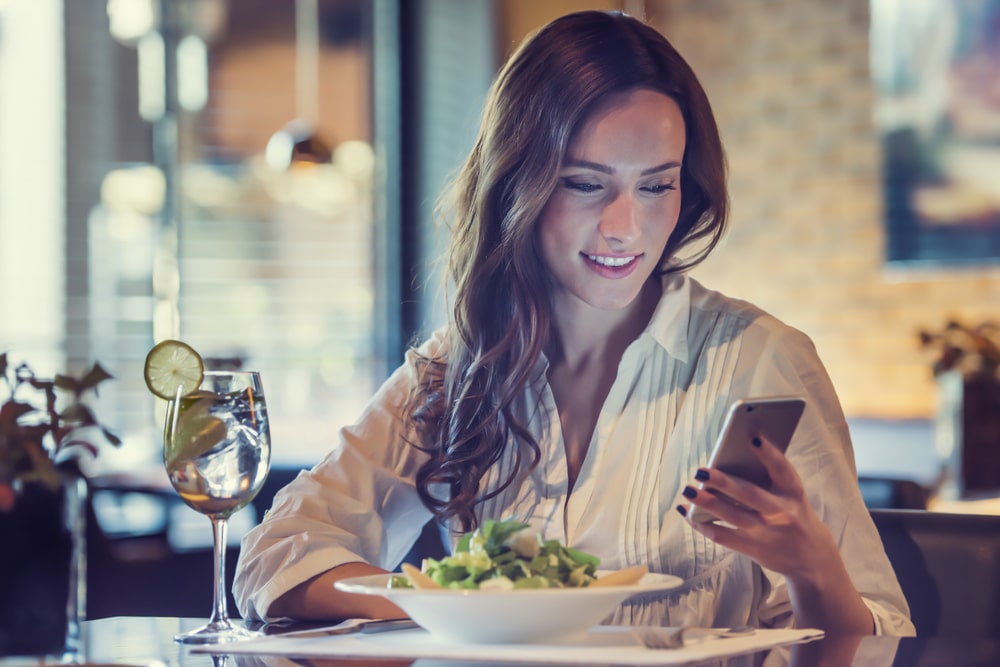 A lady enjoying a salad and a glass of water in a fancy restaurant while checking her app to see if her car is charged.