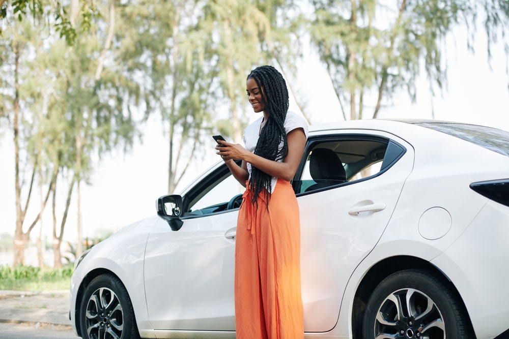 A smiling woman standing next to her car while looking at her smartphone.