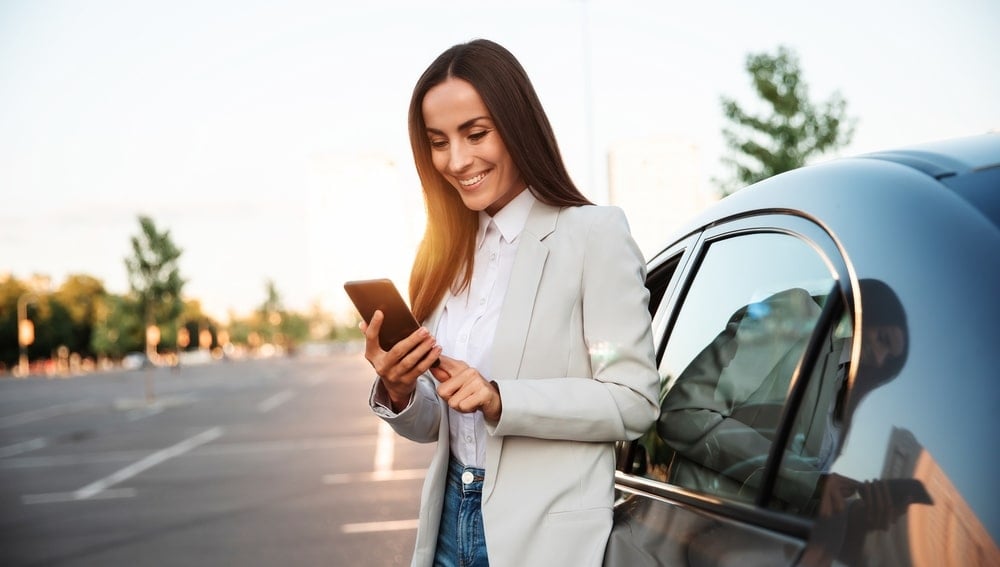 A smiling woman standing next to her electric car as she is holding her smart phone.