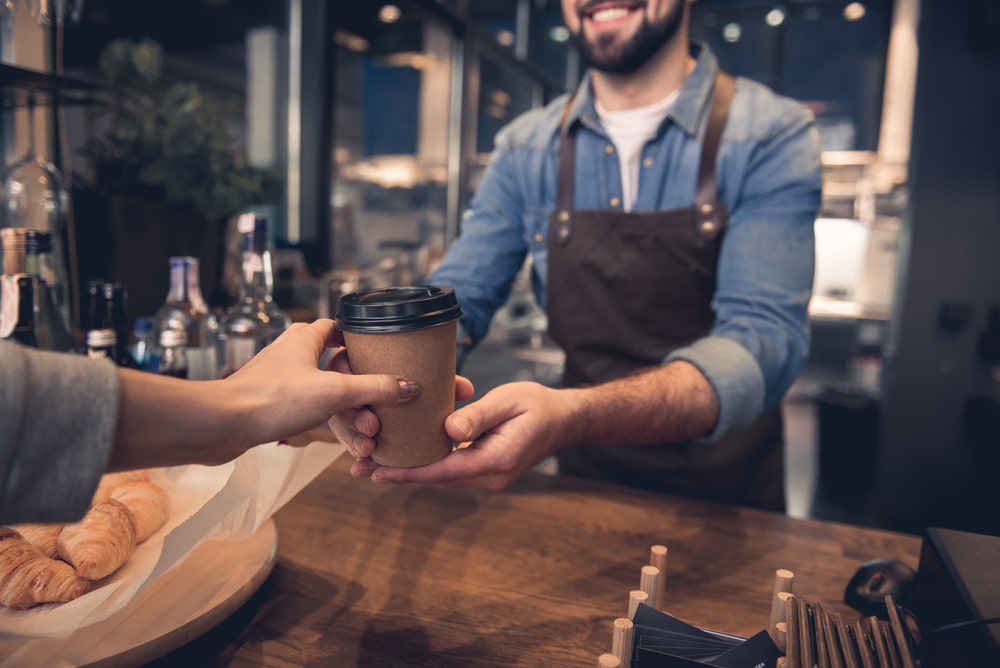 A barista handing over a coffee-to-go cup to a customer who's waiting on their car to be charged.