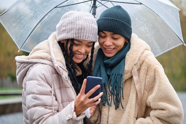 Zwei Frauen stehen unter einem Regenschirm und schauen auf ein Smartphone.