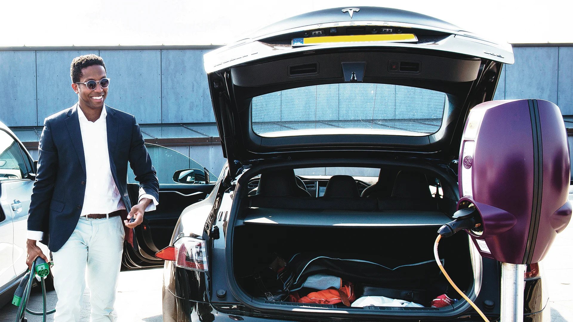 A man with a charging cable in his hand walks next to his black EV with the trunk open, towards a purple EVBox BusinessLine charging station.