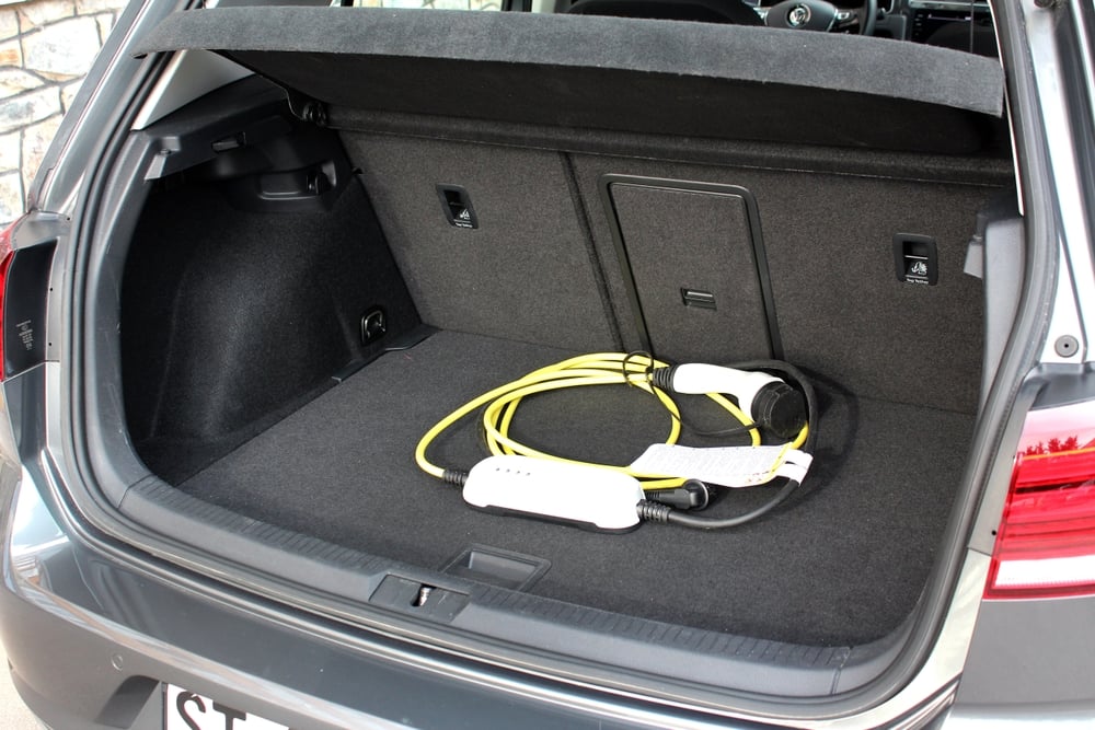 A yellow charging cable with a white charging plug sits in an open trunk of a dark grey EV