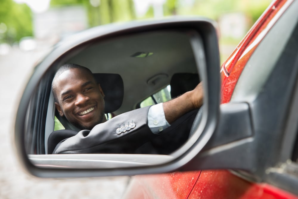 A side-mirror view of a male driver smiling in car.
