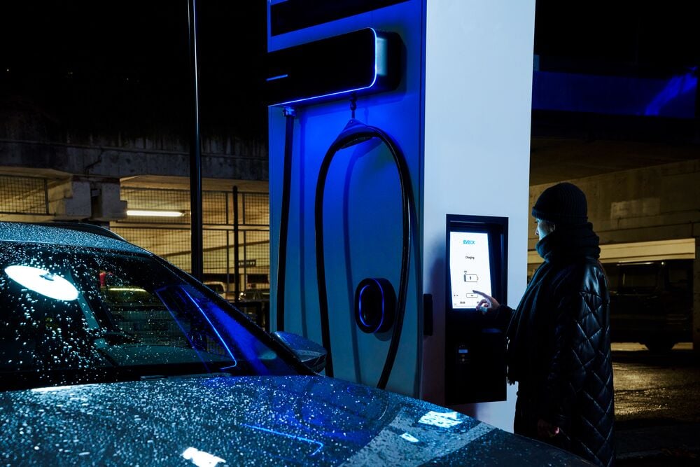 A man charges his EV using an EVBox Troniq Modular charging station in the rain at night.