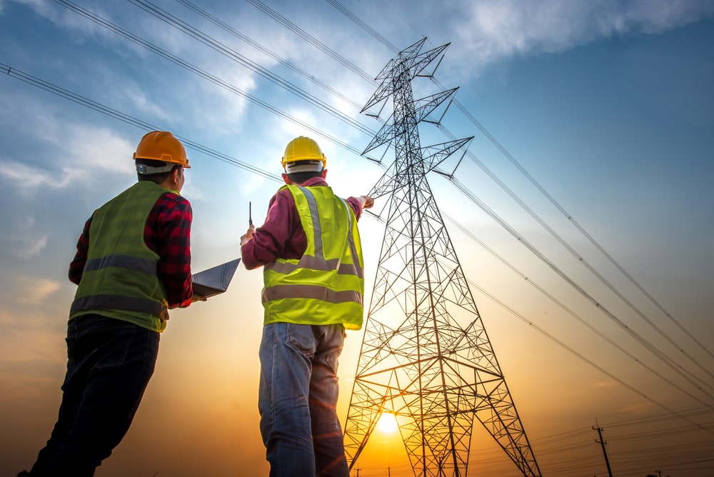Two men in construction vests and hats pointing up at at a electric power tower