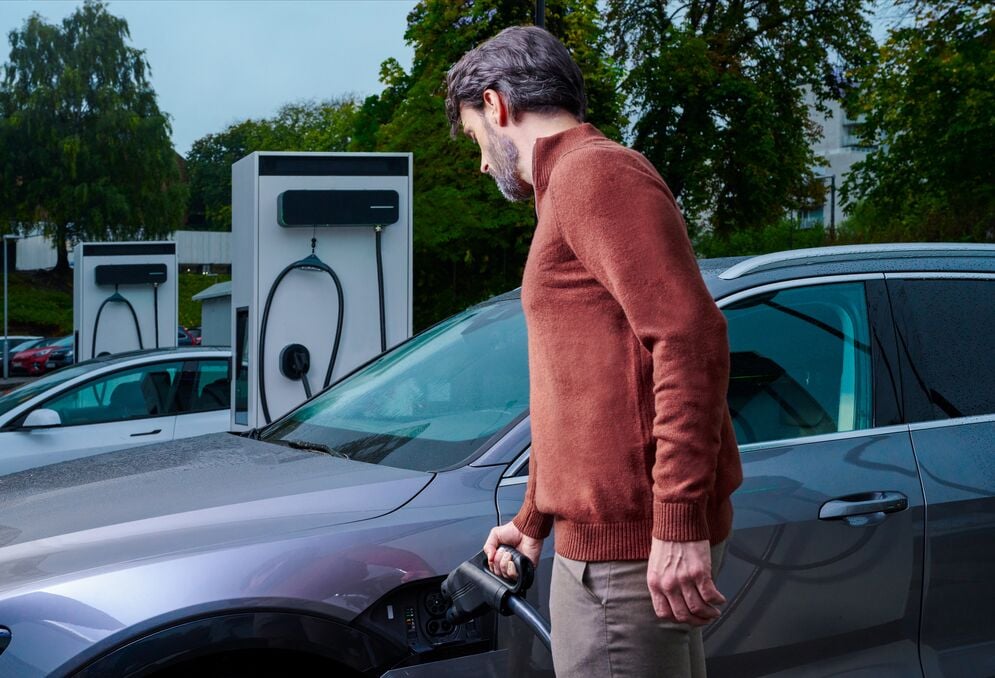 A man wearing a red sweater and khaki pants plugging in the DC connector to his vehicle to use a public EVBox Troniq Modular fast charging station to charge his car.