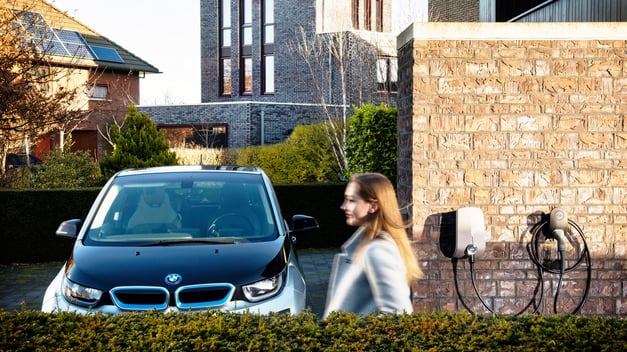 Eine Frau geht in einem Wohngebiet an einem Elektro-BMW und einer EVBox Elvi Heimladestation vorbei.