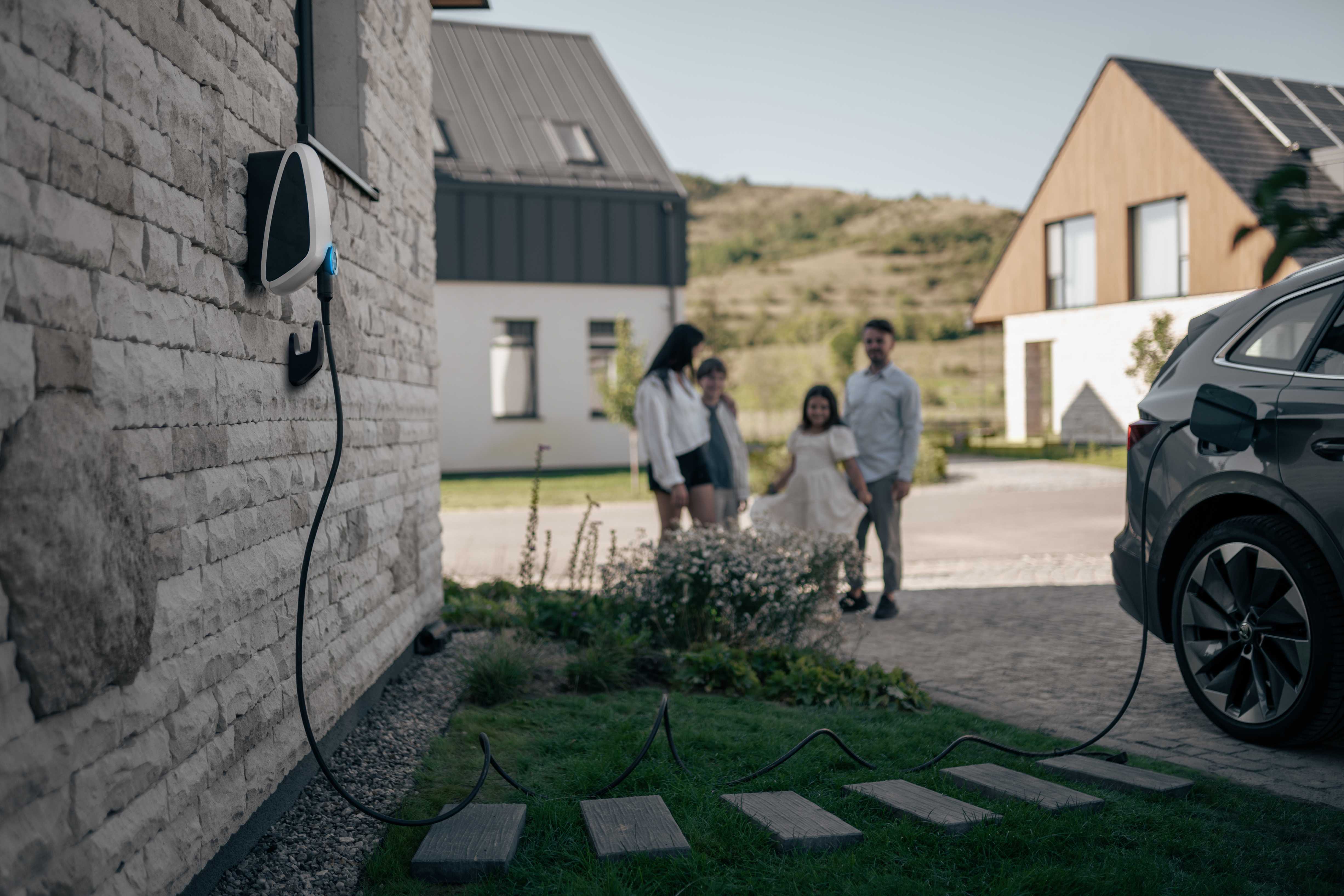 An Elvi charger mounted on the house's brick wall while the EV is connected to it. At the background, a family is enjoying their time together.