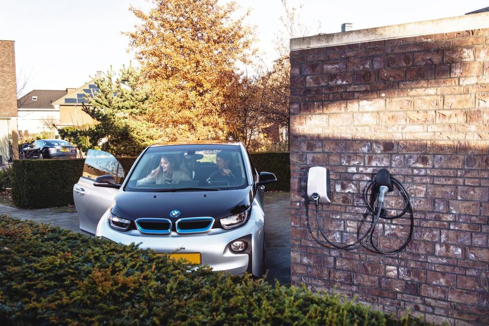 A woman is parking her electric car in the driveway, while another woman is getting out of the car. On the right, an EVBox Elvi home charging station is mounted on a wall and ready to charge.