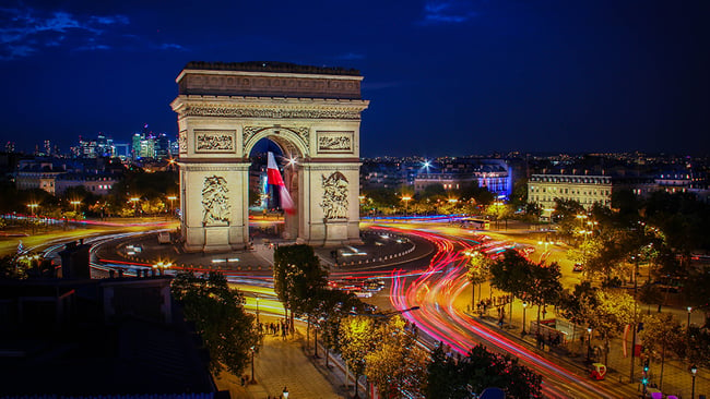 An aerial shot of the Arc de Triomphe in Paris, France at night. The roads are filled with streaks of lights coming from passing cars.