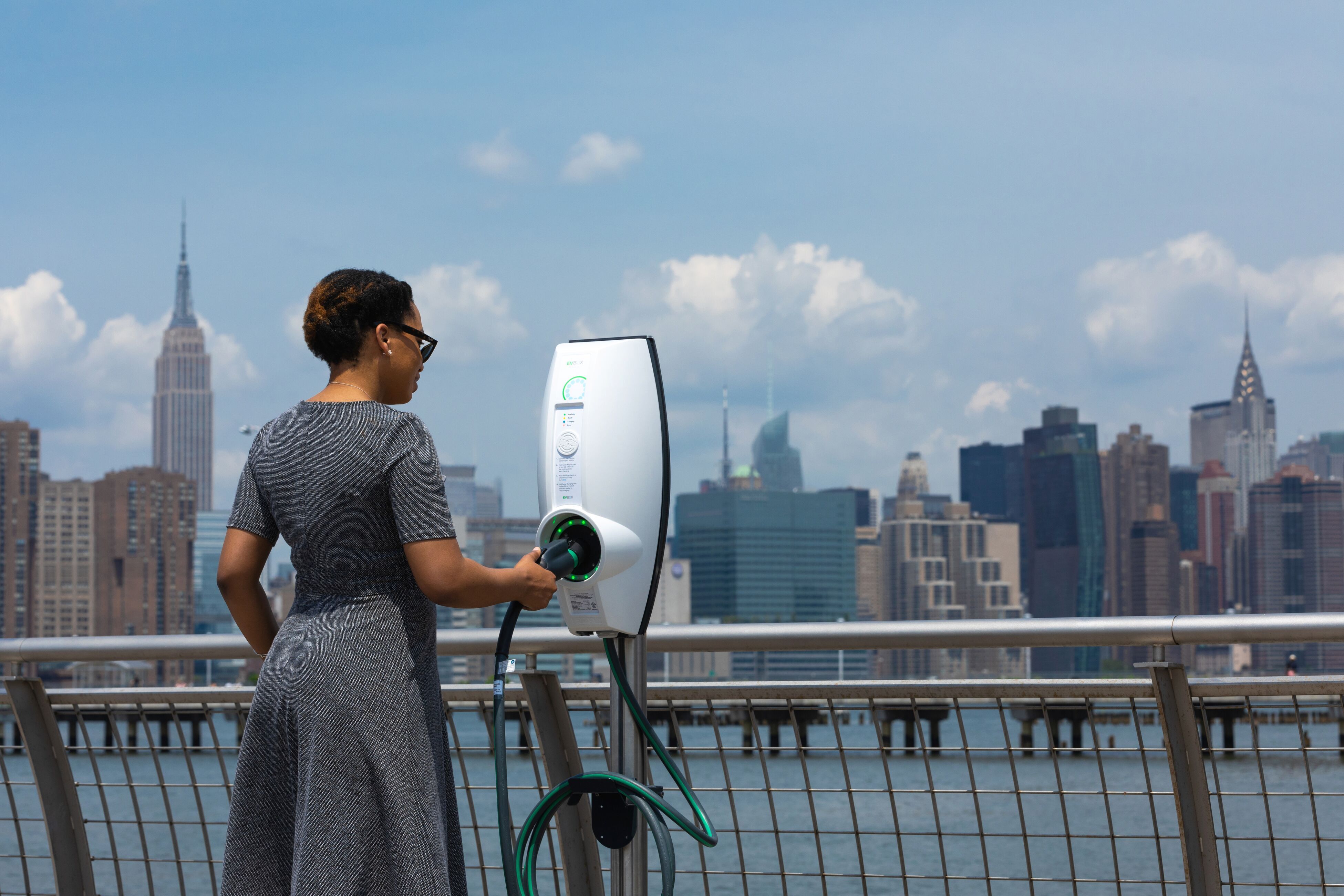 A woman is holding the charging plug of a white EVBox charging station. In the background, the cityscape of New York on a sunny day.