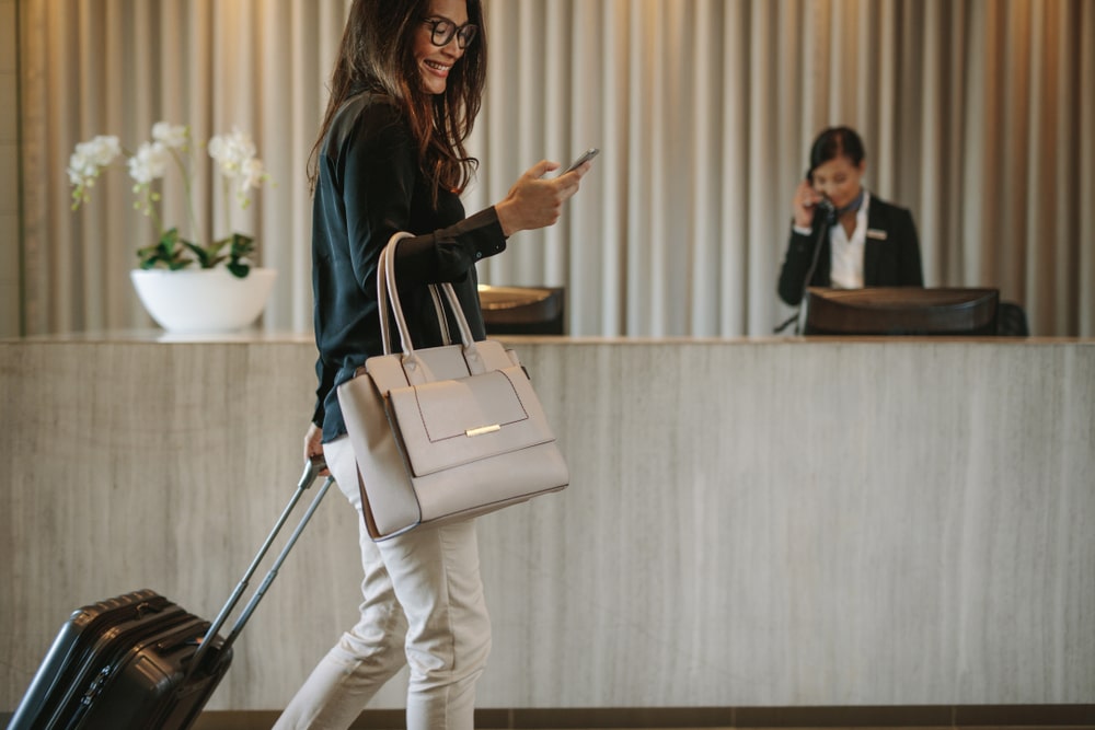 Woman carrying suitcase and a purse, smiling while looking at her cell phone, walks by the lobby of an upscale hotel.