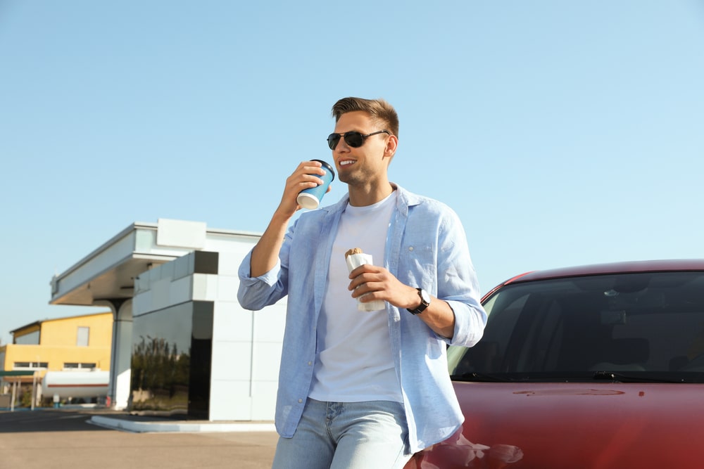 Man wearing sunglasses leaning against red car drinks from coffee cup and holds a sandwich.