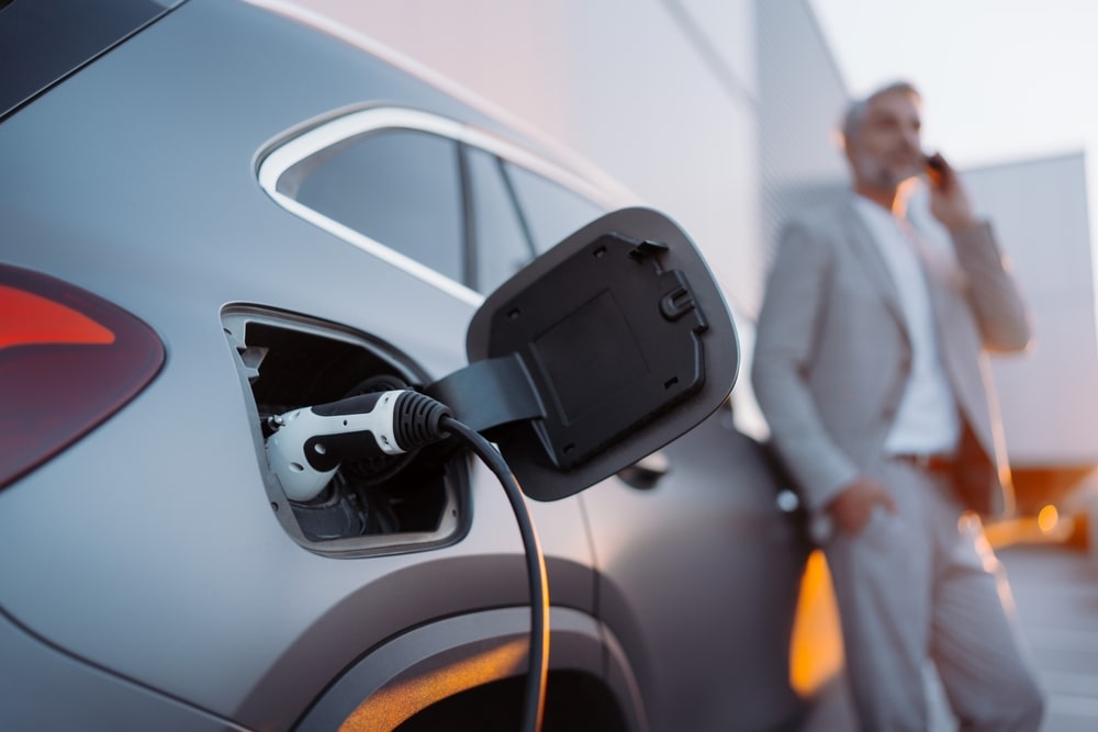 Man in suit leaning against electric car being charged whilst talking on the phone.