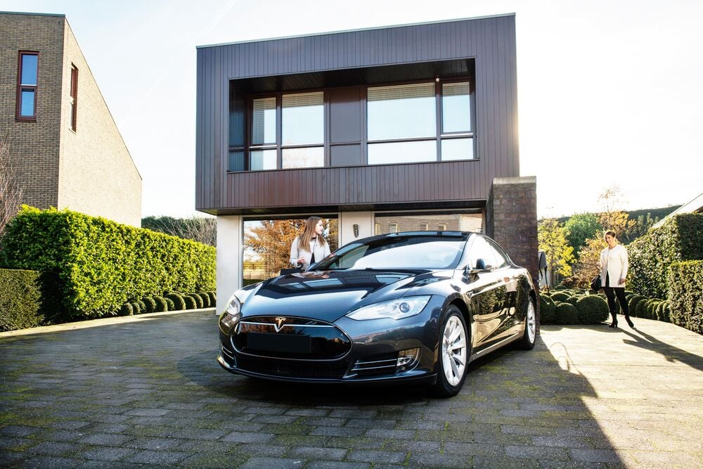 A black electric car is parked in front of a modern house on a sunny day. A woman in approaching the vehicle and another person is standing in the background, next to the house.