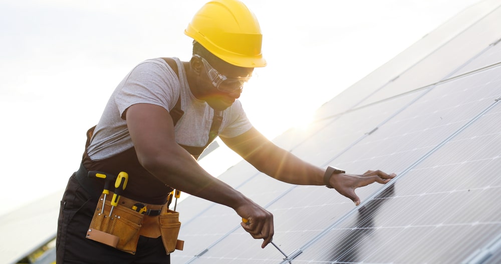 Ein Handwerker mit Helm und Werkzeuggürtel installiert Solarpaneele auf einem Dach.