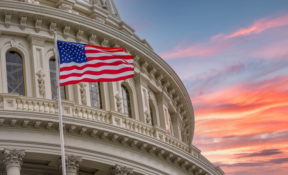 View of the United States Capitol Rotunda Dome in Washington DC with the Star Spangled American Flag against colorful dramatic sunset sky background.