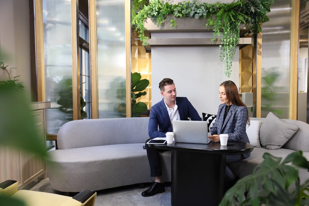 Two colleagues sitting down on a comfortable grey couch in an office space surrounded by plants.