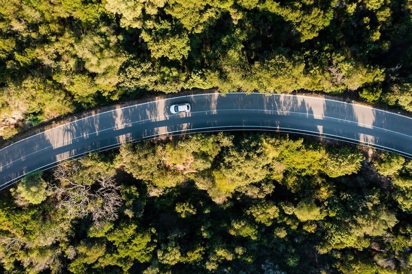 An areal shot of a car driving on a highway road in the middle of a forest area. 