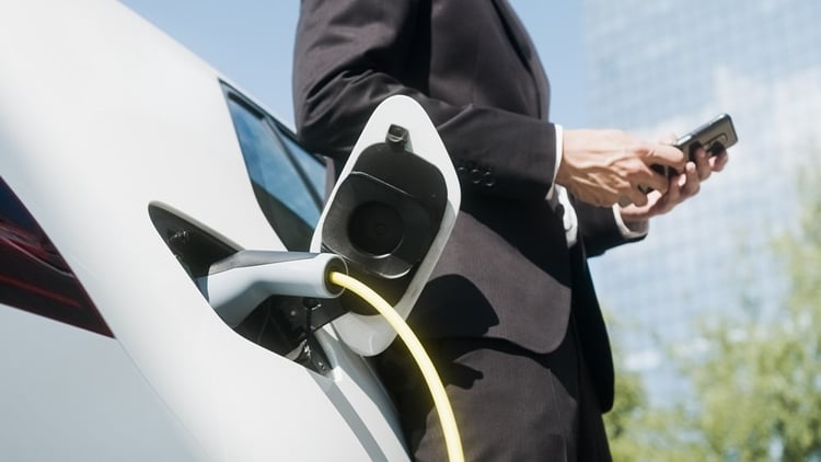 Mann in schwarzem Anzug lädt sein Elektroauto auf, lehnt sich dagegen und hält sein Smartphone in der Hand.