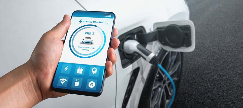 A close-up of a hand holding a smartphone and showing data about an electric car's EV charging session charging in the background.