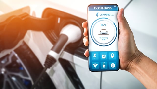 A hand holding a phone showing the charging percentage of the electric car in the background.