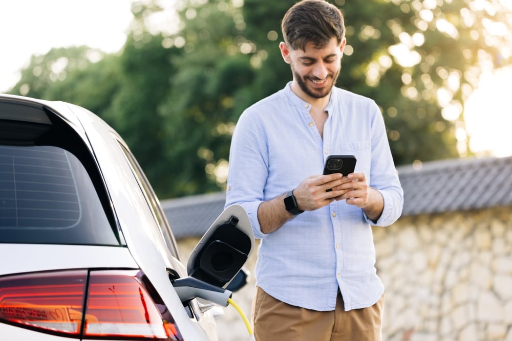 Een man lacht naar zijn telefoon terwijl hij naast zijn elektrische auto staat die aan het opladen is.