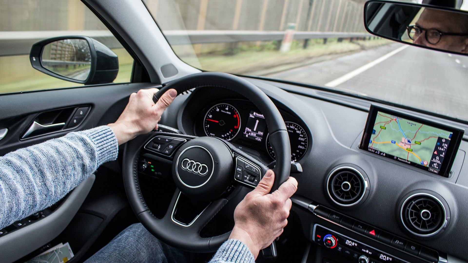 A middle aged man in a sweater driving an Audi in what looks like a moderately cloudy place.
