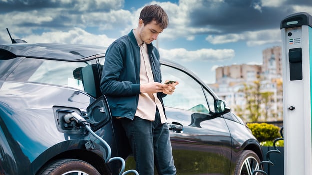 Ein moderner Mann lehnt sich an sein Elektroauto und vertreibt sich die Zeit mit seinem Handy, während das Auto aufgeladen wird.