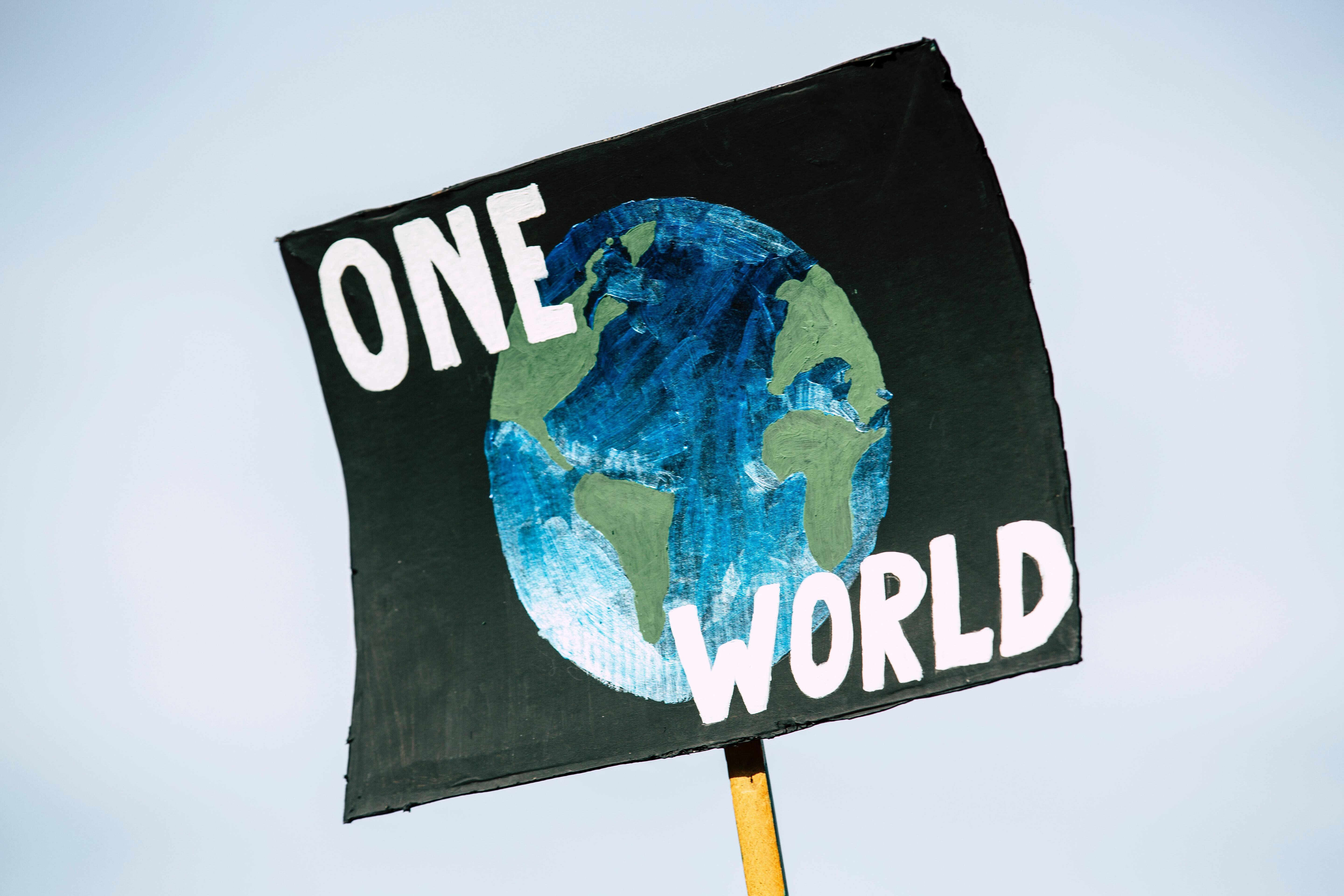 A close-up shot of a black protest sign that shows a drawn image of planet earth accompanied by bold white-painted letters that read: ONE WORLD.