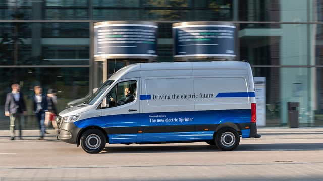 A Mercedes eSprinter is driving fast in a urban environment. There are some defocused people walking on a sidewalk behind the van.