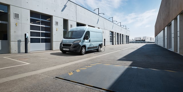 A Peugeot e-Boxer is parked and charging outside some warehouses on a sunny day.