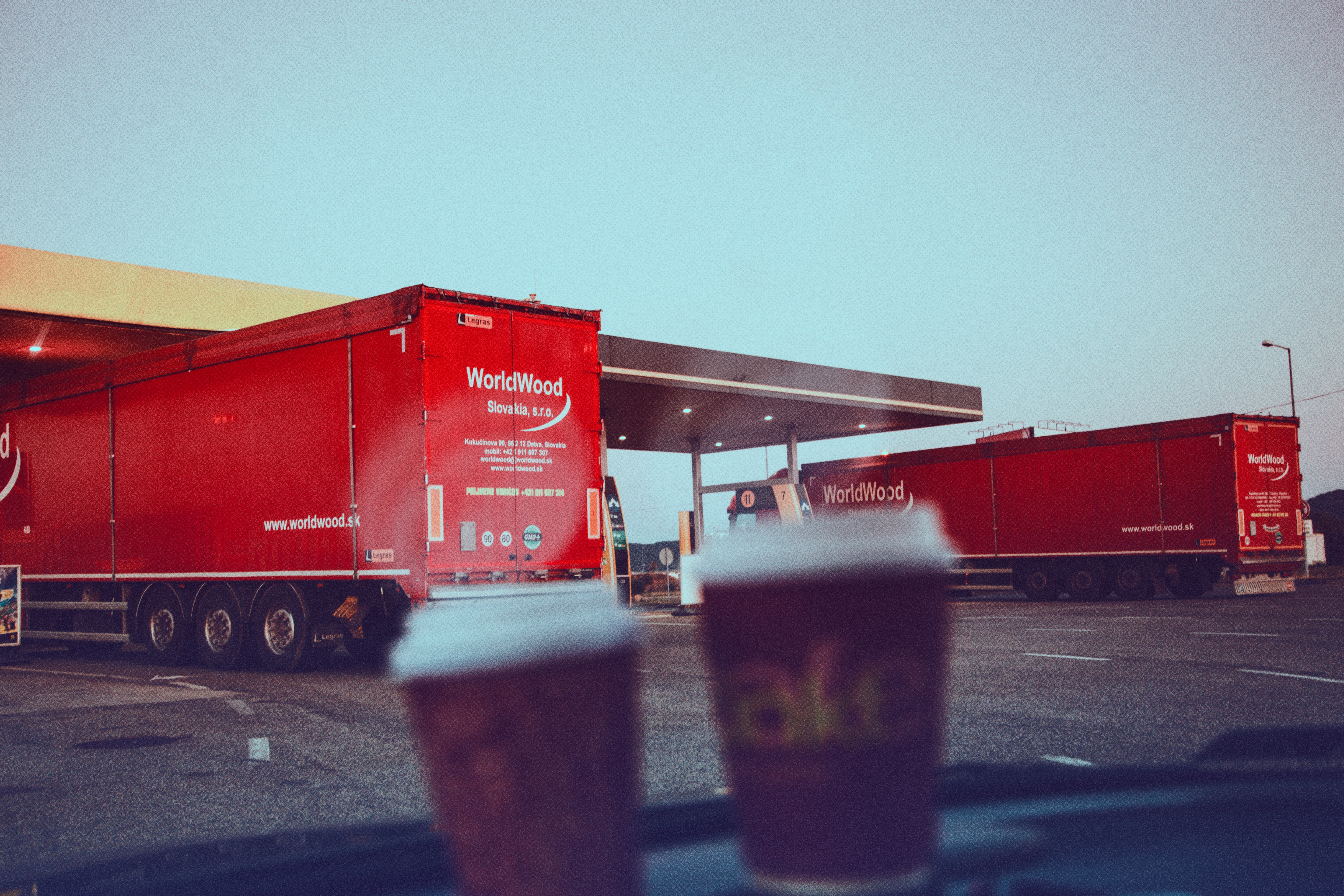 Photo taken from the inside of a vehicle, where two take-away coffee cups rest on the dashboard. through the windshield we see  two heavy-duty trucks taking a break mid route at a fuel retail location.