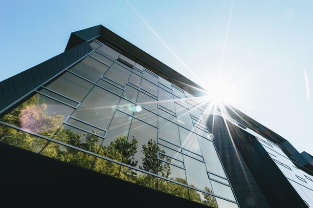 The exterior of an office building breaking the sunlight and showing the reflection of lush trees in the windows.
