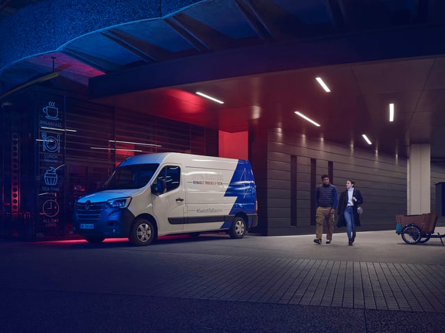 A Renault Master E-Tech is parked outside an enlightened cafeteria at night. A couple is walking by close to the vehicle.