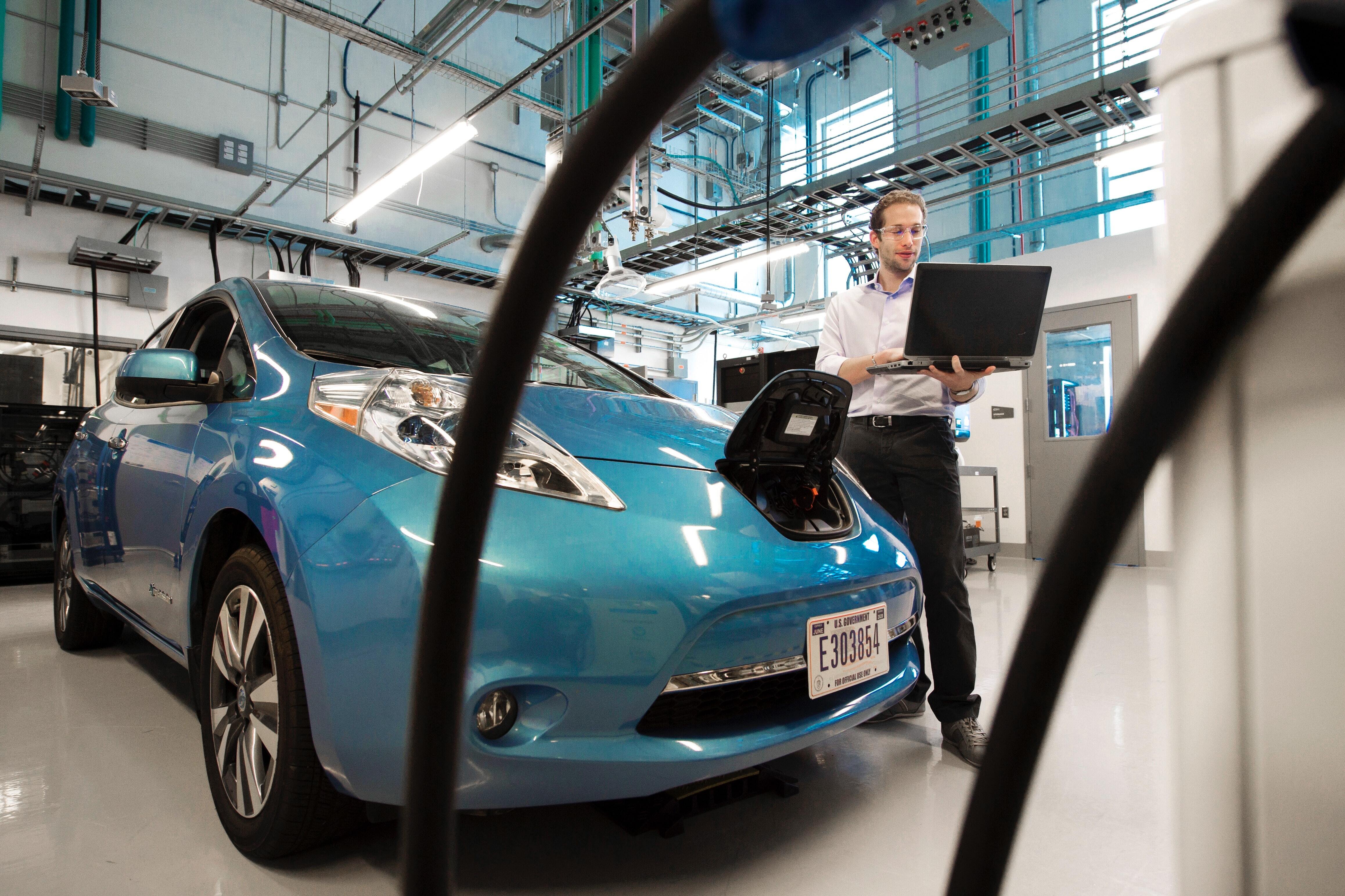 A man holding a laptop next to an EV, seemingly checking the status of the battery.
