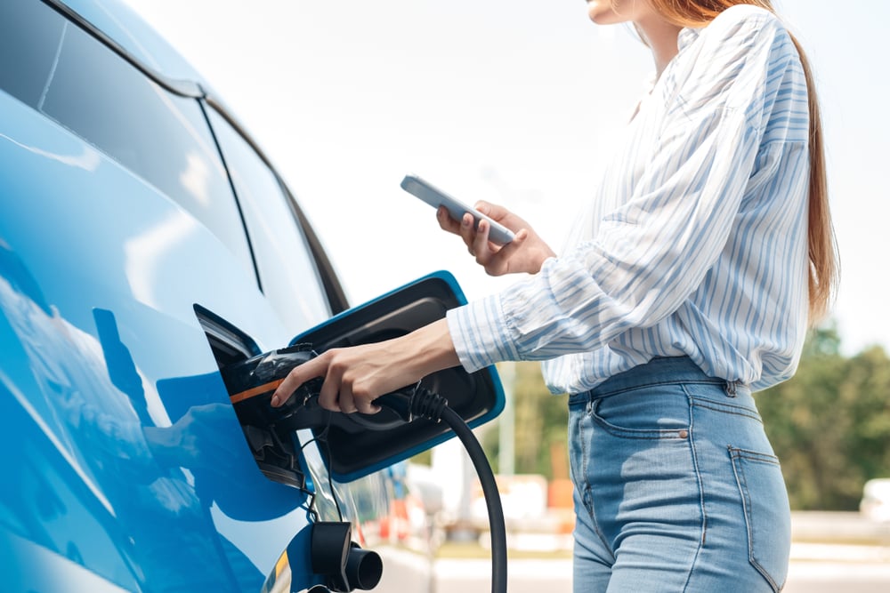 A closeup of a woman connecting the charging plug to an electric car while looking at her smartphone.