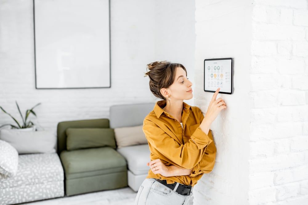 Eine Frau lehnt an einer Wand im Inneren eines Hauses. Sie stellt einige smarte Funktionen ein, um die Geräte in ihrem Haus mit einem Tablet zu steuern.
