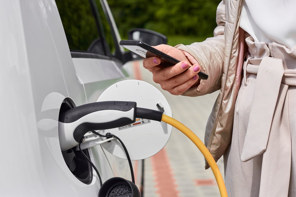 A closeup of a woman plugging her electric car to charge it, while holding her smartphone on the other hand.