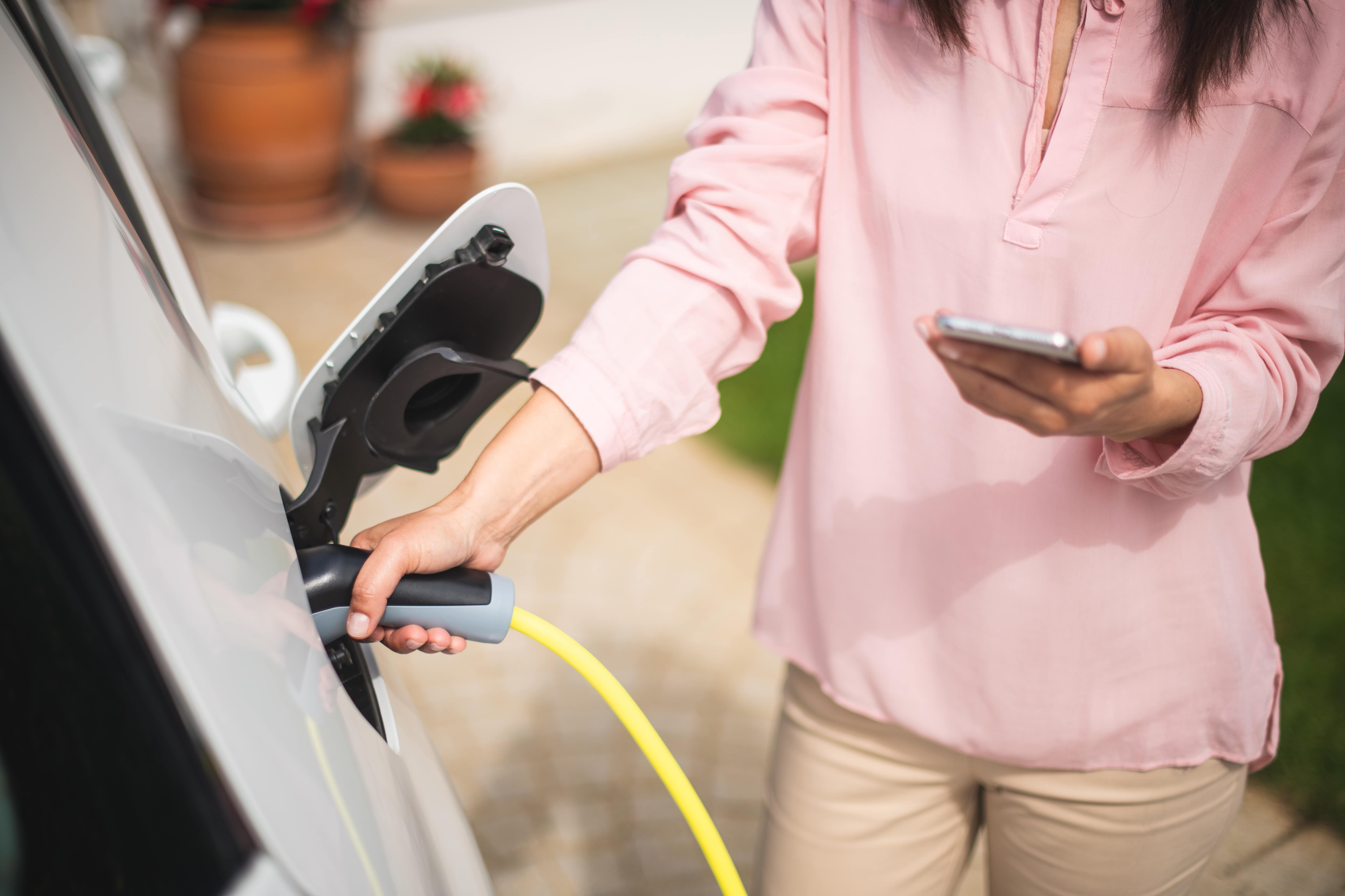 A woman, holding an electric car charging cable that's plugged into her vehicle on her driveway in one hand, whilst she casually holds her smart phone with the other.