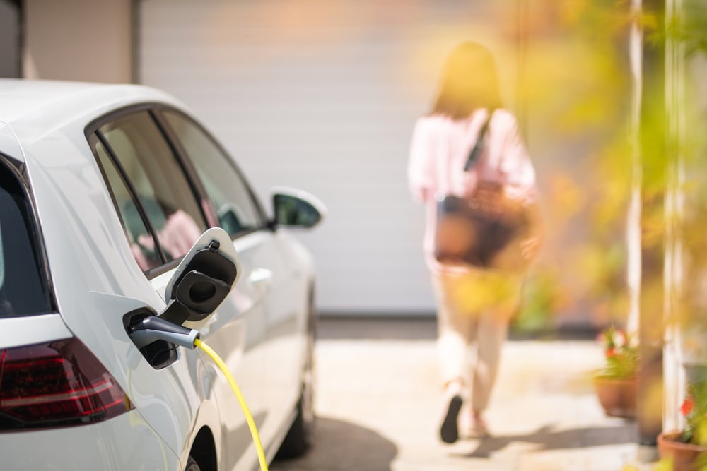Een elektrische auto laadt op in de zon op de oprit voor een garage. Op de achtergrond loopt een vrouw met tas rustig weg naar de voordeur van haar huis